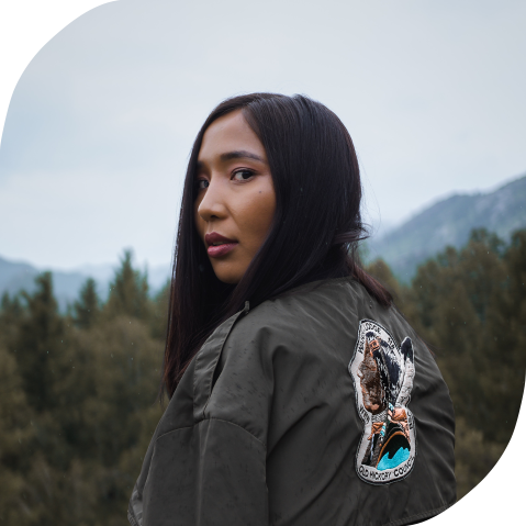 A young Indigenous woman glances over her shoulder on a cloudy day. Behind her are mountains and a forest.